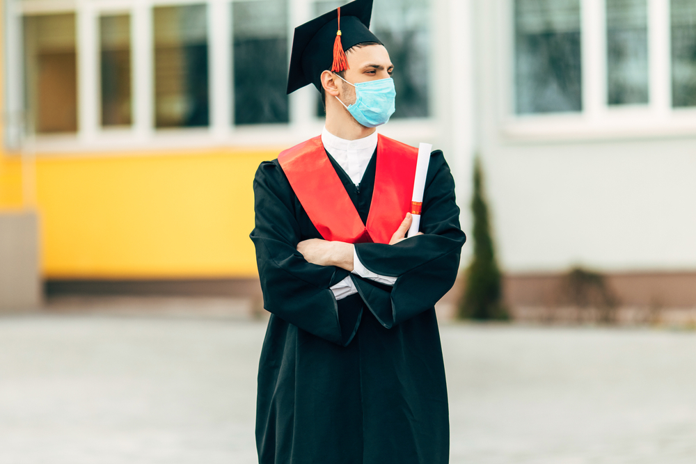 A male graduate student wears a protective mask against coronavirus, in a black graduation dress, with a diploma in his hands. Concept of graduation ceremony, quarantine, coronavirus
