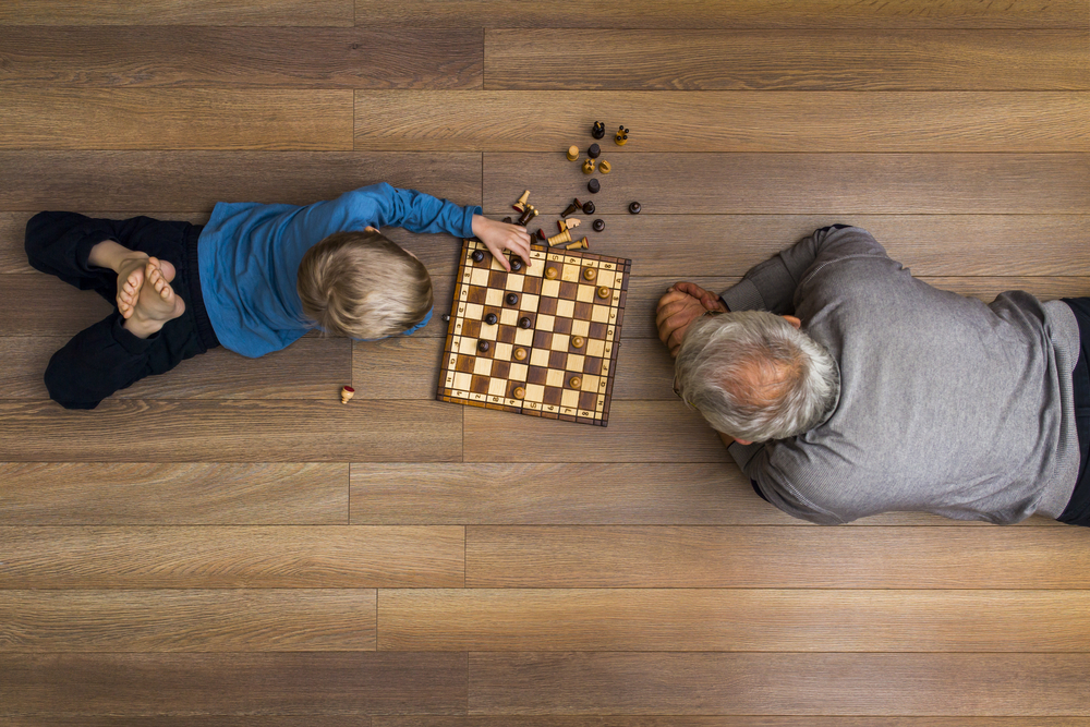 Small boy laying on the wooden floor and playing chess with his grandfather after setting holiday sleeping arrangements