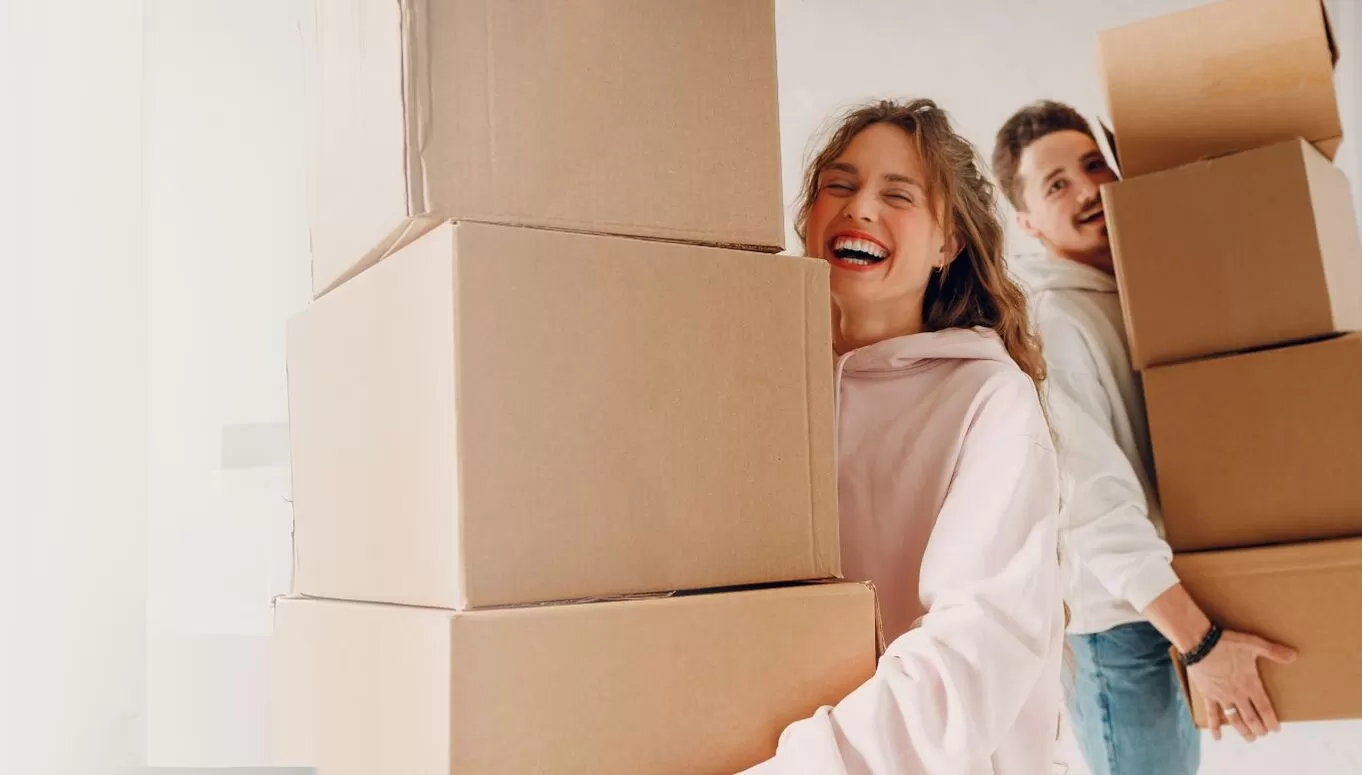A joyful woman carrying a stack of cardboard boxes with a man behind her, both engaged in moving or packing activities.