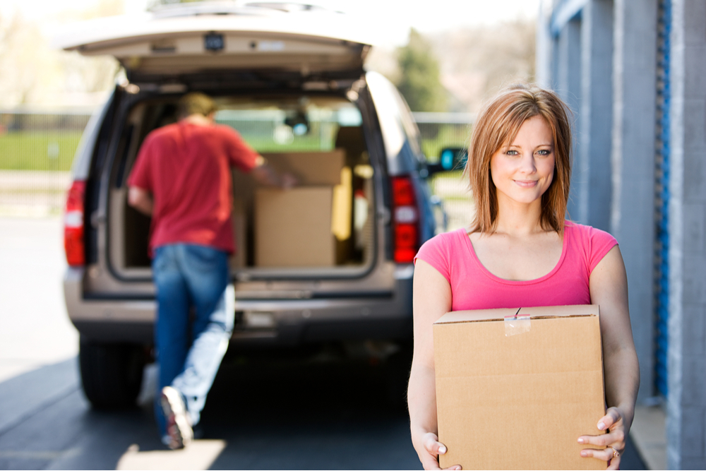 Woman in pink shirt smiling and carrying a moving box outside a storage facility, with a vehicle being loaded in the backdrop.
